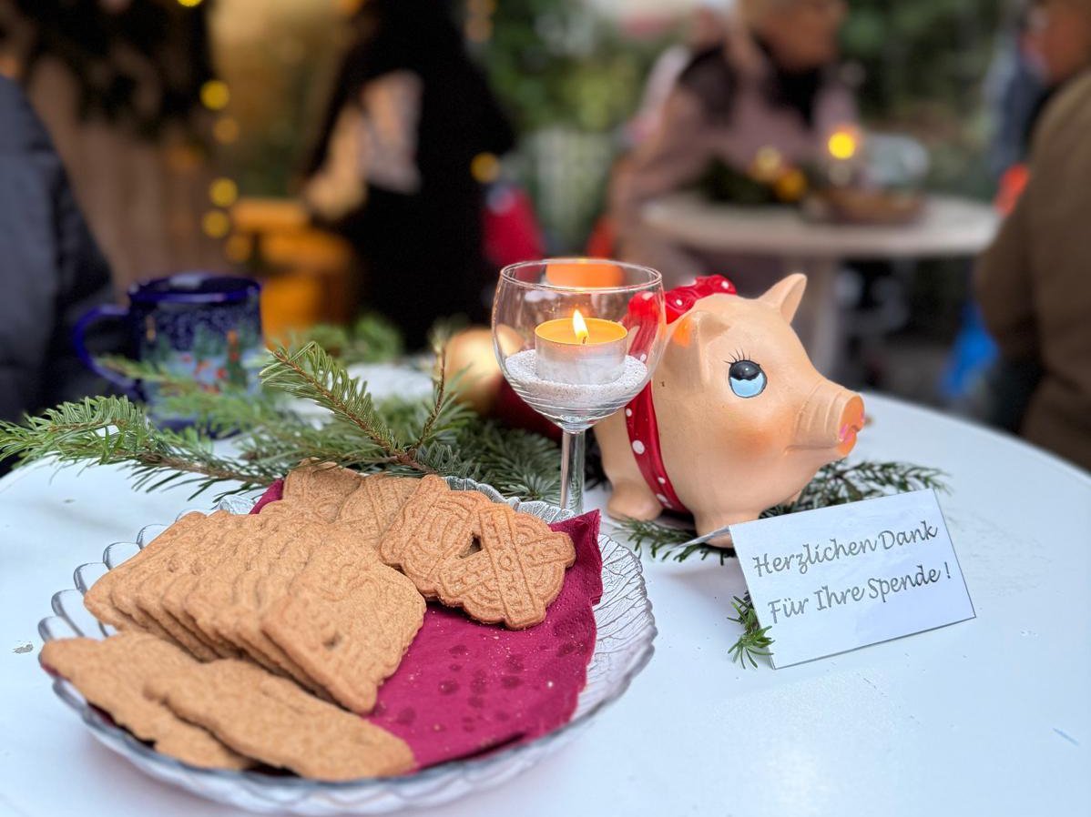 Auf einem runden Stehtisch stehen weihnachtlich dekorierte Kleinigkeiten: ein Teller mit Spekulatius-Keksen, ein kleines Glas mit brennendem Teelicht, Tannenzweige und ein rosafarbenes Sparschwein mit rotem Schal. Daneben liegt ein Schild mit der Aufschrift „Herzlichen Dank für Ihre Spende!“. Im unscharfen Hintergrund sitzen und stehen Menschen unter Lichterketten, was eine gemütliche Weihnachtsmarkt-Atmosphäre vermittelt.
