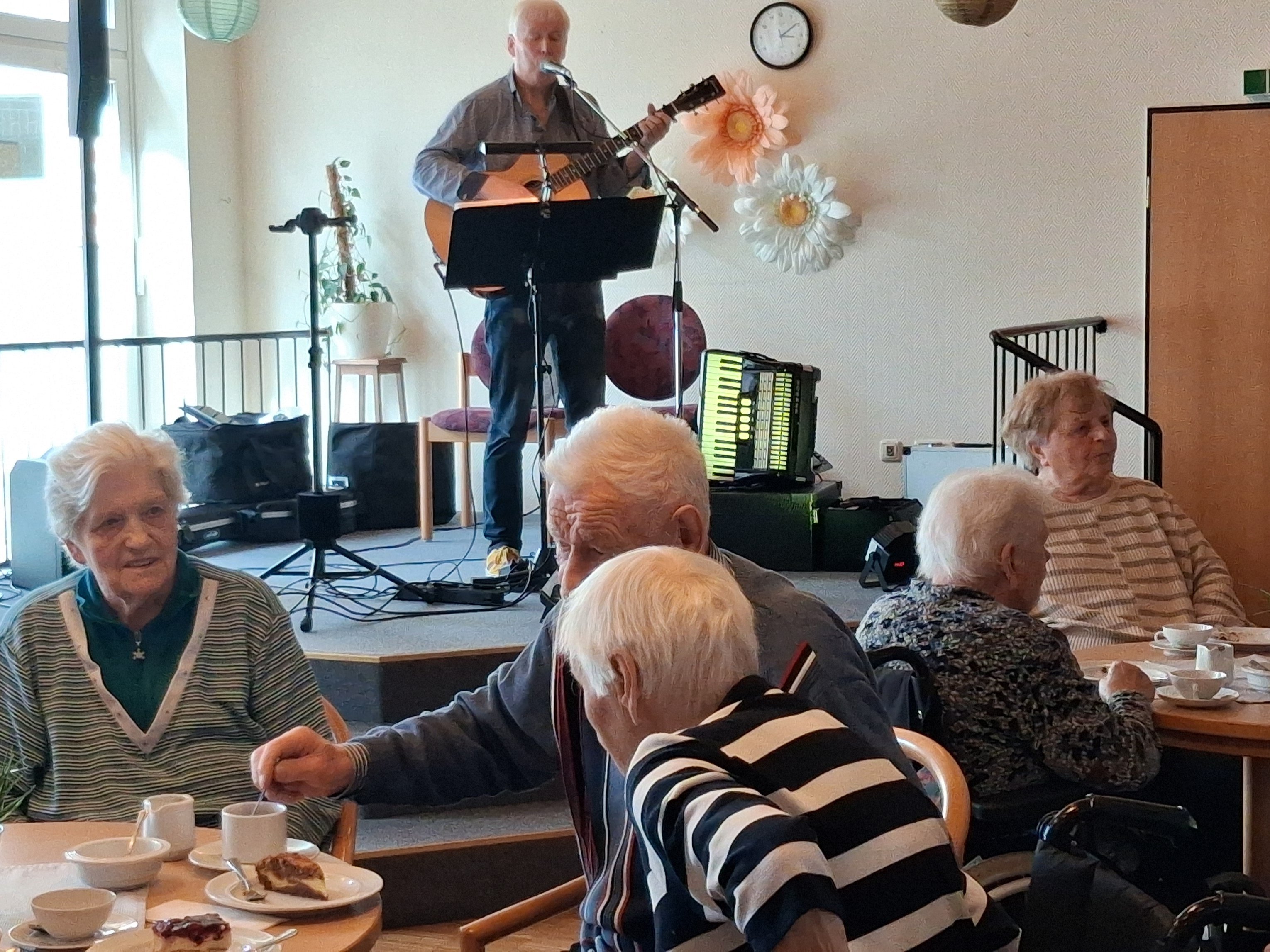 Musiker Peter Focken singt auf einer kleinen Bühne und spielt Gitarre, während Seniorinnen und Senioren an gedeckten Kaffeetischen sitzen.