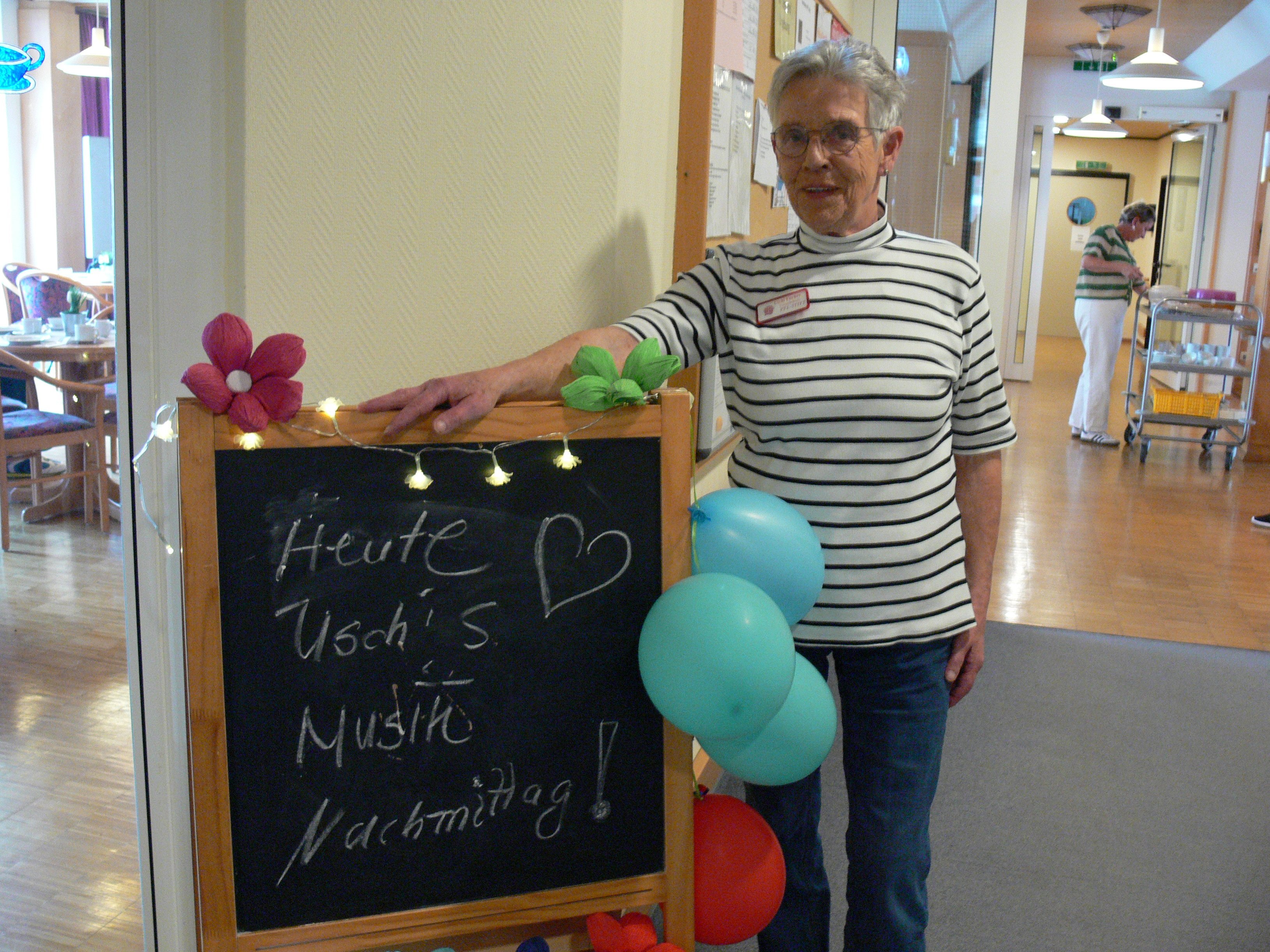 Eine Mitarbeiterin steht neben einer mit Blumen, Lichterkette und Luftballons geschmückten Tafel mit der Aufschrift „Heute Uschi's Musik Nachmittag!“