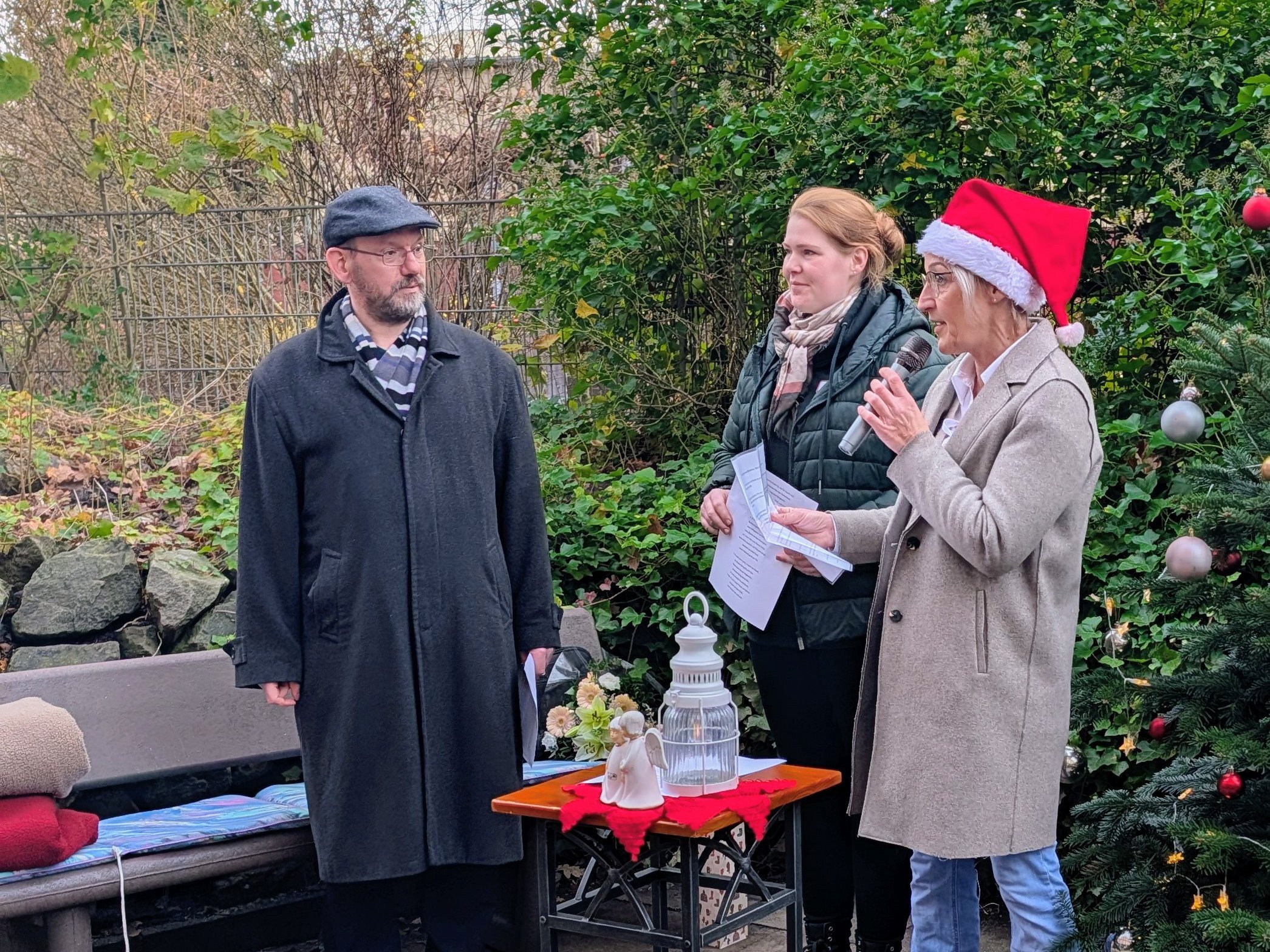 Drei Personen stehen im Freien neben einem geschmückten Weihnachtsbaum; eine Frau mit Nikolausmütze spricht ins Mikrofon. Auf einem kleinen Tisch stehen Laterne, Kerze und eine Engelsfigur.
