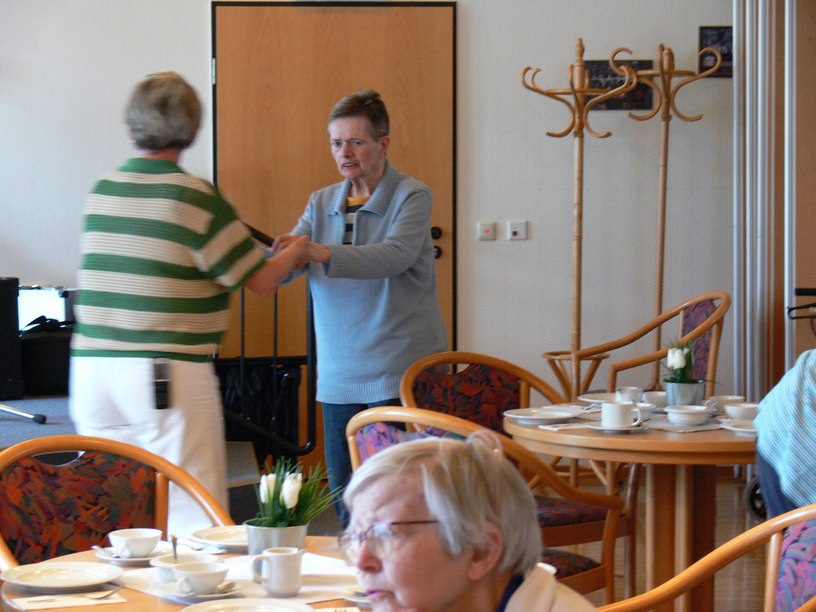 Zwei Frauen stehen sich im Gemeinschaftsraum gegenüber und halten sich beim Tanzen an den Händen. Im Vordergrund stehen gedeckte Kaffeetische.