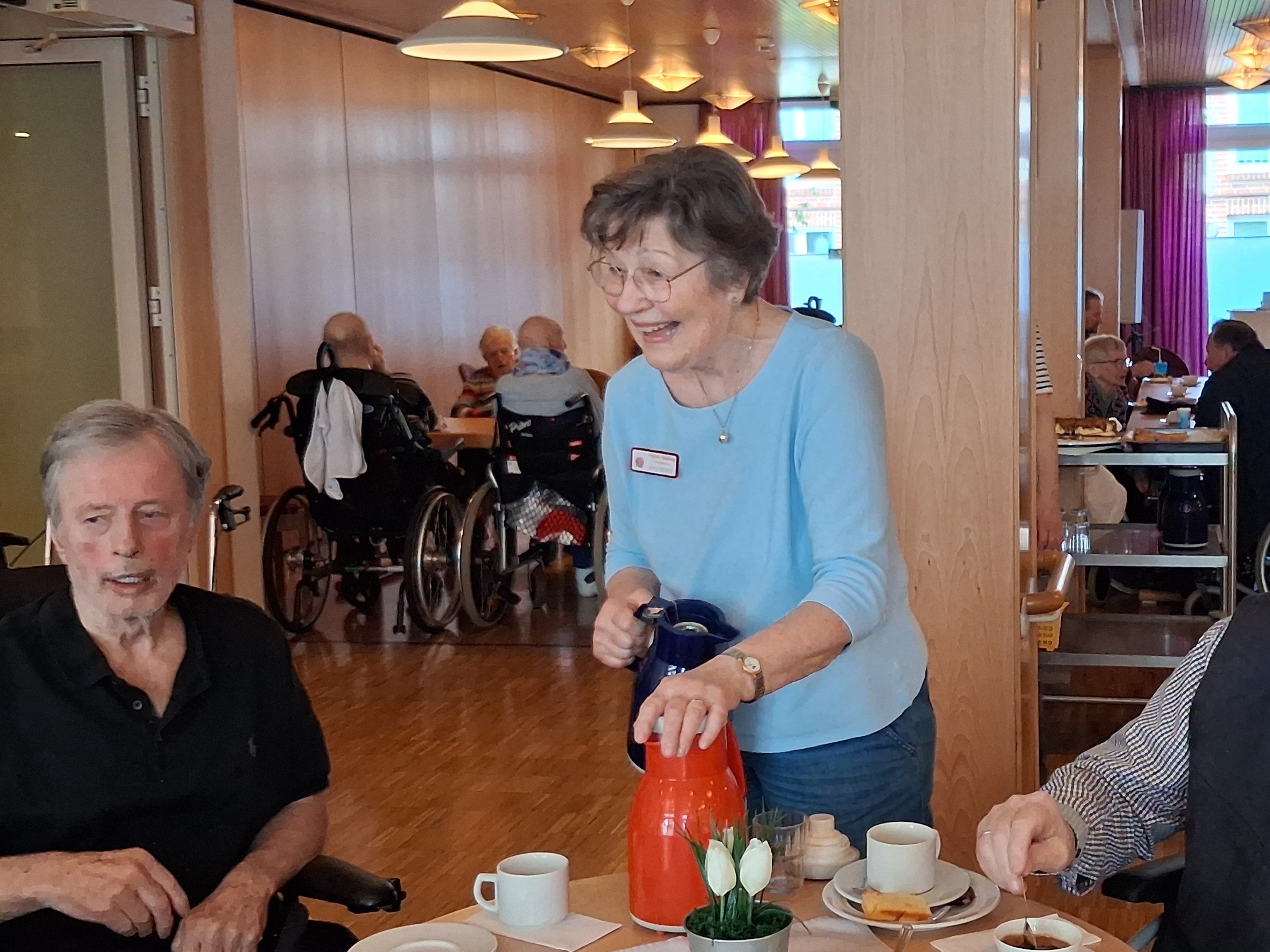 Eine lächelnde Mitarbeiterin in einem hellblauen Oberteil steht mit zwei Kaffeekannen zwischen zwei älteren Männern an einem gedeckten Tisch.