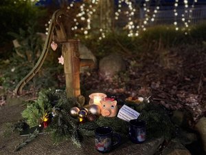 Auf einem alten steinernen Brunnen im Freien liegt eine weihnachtliche Dekoration aus Tannenzweigen, kleinen Christbaumkugeln und einer brennenden Kerze im Glas. Daneben stehen zwei dunkelblaue Tassen und ein kleines rosafarbenes Spendenschwein. Im Hintergrund leuchten Lichterketten in den Bäumen und sorgen in der Abenddämmerung für eine ruhige, festliche Atmosphäre.