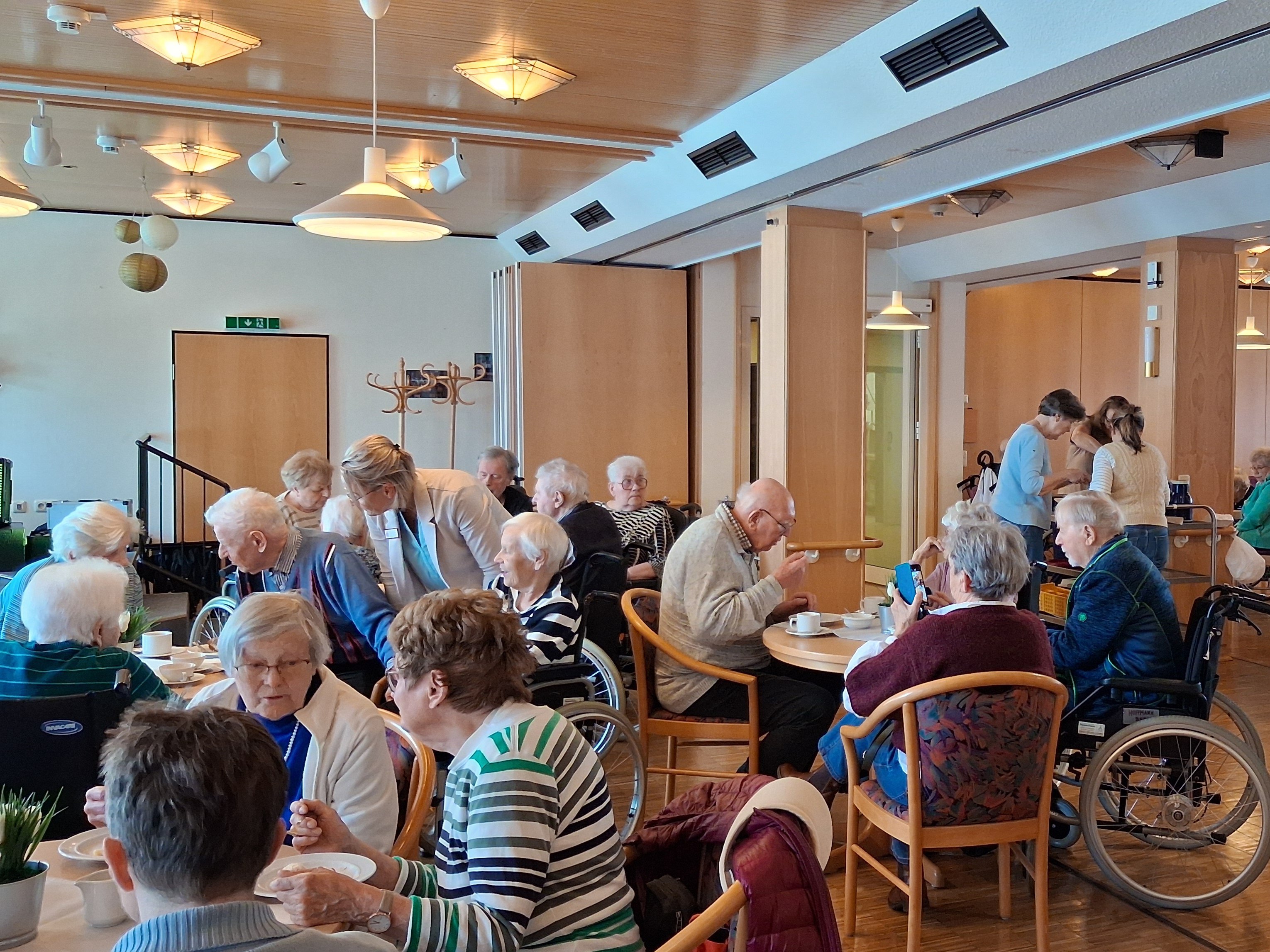 Seniorinnen und Senioren sitzen in kleinen Gruppen an gedeckten Tischen. Mehrere Mitarbeitende sind im Raum unterwegs und unterhalten sich mit den Gästen.
