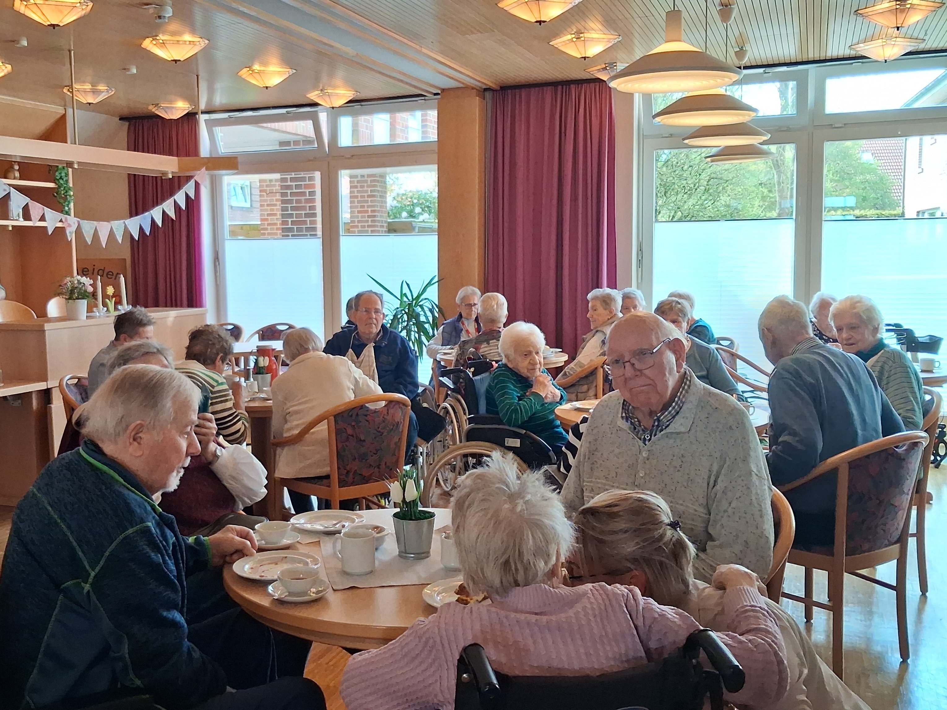 Zahlreiche Seniorinnen und Senioren sitzen an mehreren runden Kaffeetischen in einem hellen Gemeinschaftsraum und unterhalten sich.