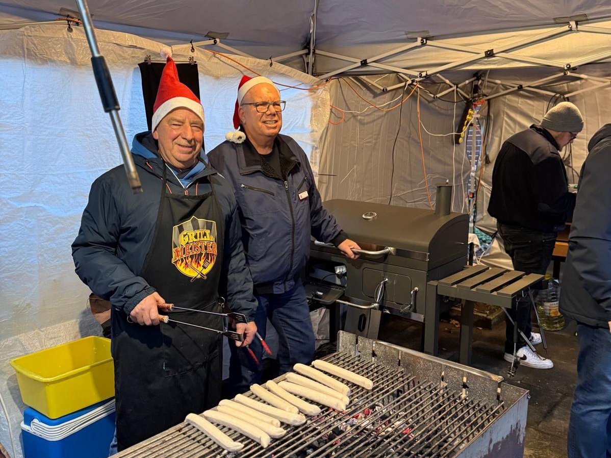 Mit Nikolausmützen und guter Laune stehen zwei Ehrenamtliche in einem Pavillon am Grill: Vor ihnen brutzeln mehrere helle Würstchen auf dem Rost, daneben steht ein großer Grillwagen. Im Hintergrund arbeiten weitere Helfer.